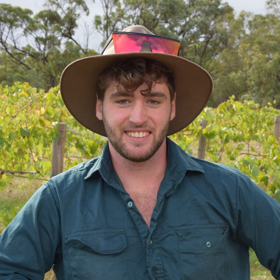 A young man with a beard and tousled hair, wearing a wide-brimmed hat with sunglasses on top and a teal shirt, stands outdoors in front of grapevines and trees, smiling at the camera.