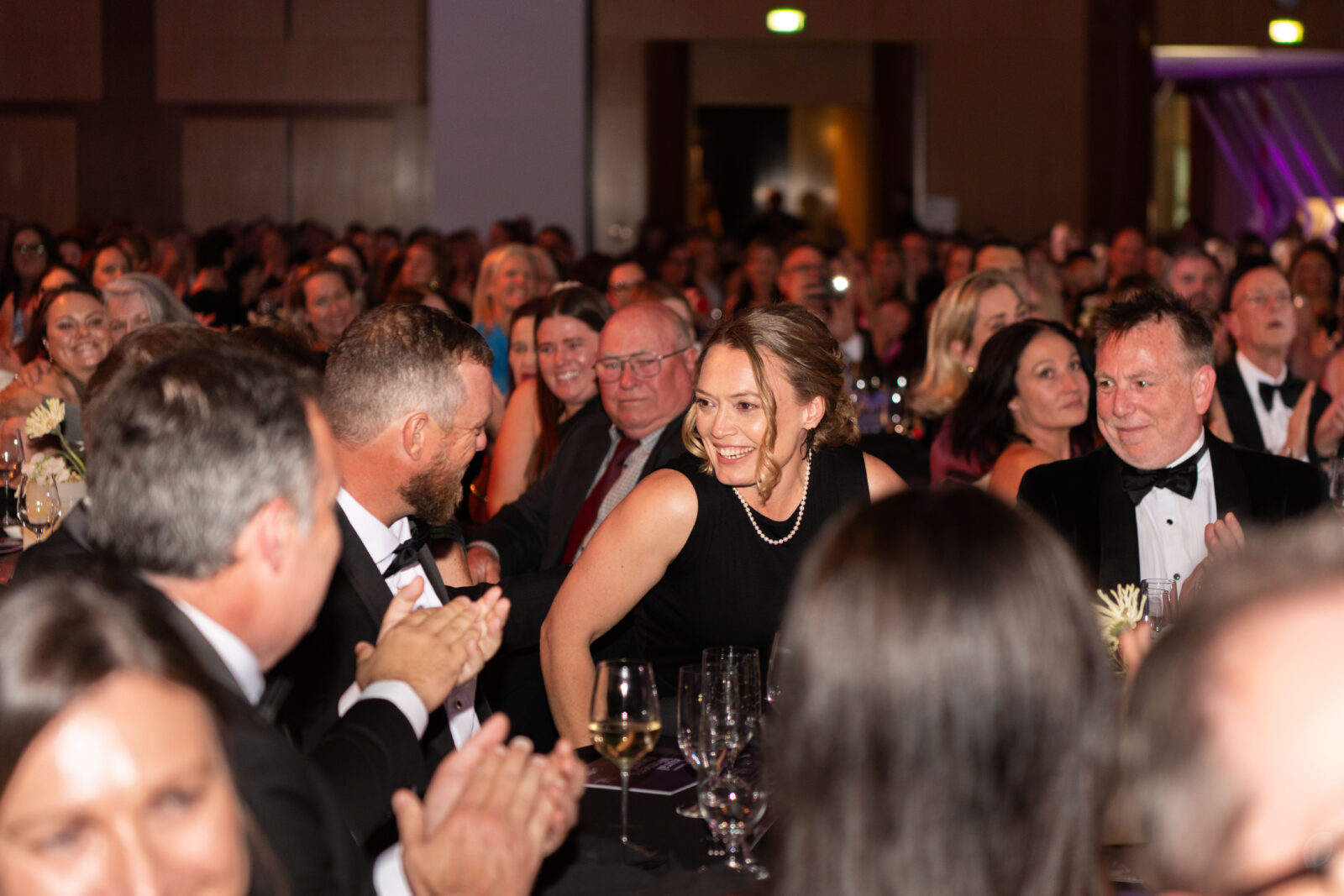 A woman in a black dress smiles whilst surrounded by applauding guests at a formal event; the room, filled with seated people in evening attire, buzzes with excitement for the highly anticipated RWA Tickets.