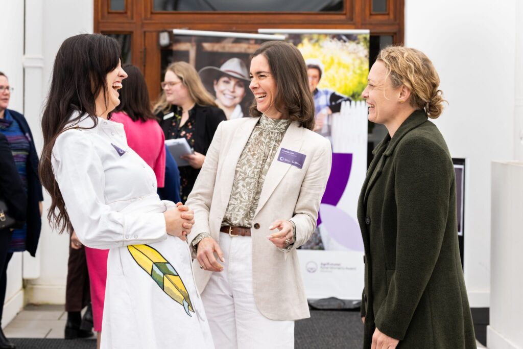 Three women stand outside, smiling and laughing together at an event. One woman wears a white dress, another a cream blazer, and the third a green coat. Other people and event signage are visible in the background.