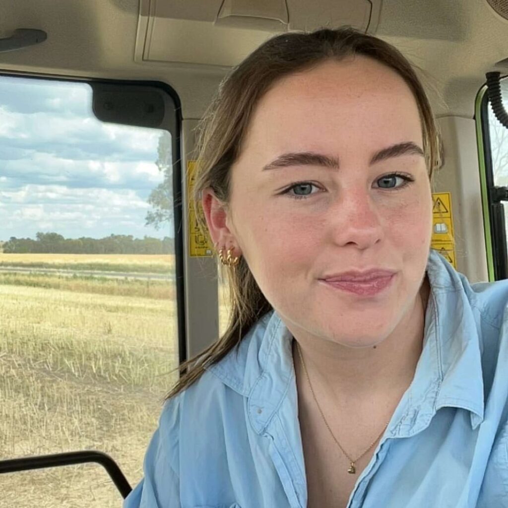 A young woman with light brown hair and blue eyes is smiling inside a vehicle with a field and blue sky visible through the window behind her. She is wearing a light blue shirt and gold jewellery.