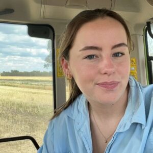 A young woman with light brown hair and blue eyes is smiling inside a vehicle with a field and blue sky visible through the window behind her. She is wearing a light blue shirt and gold jewellery.
