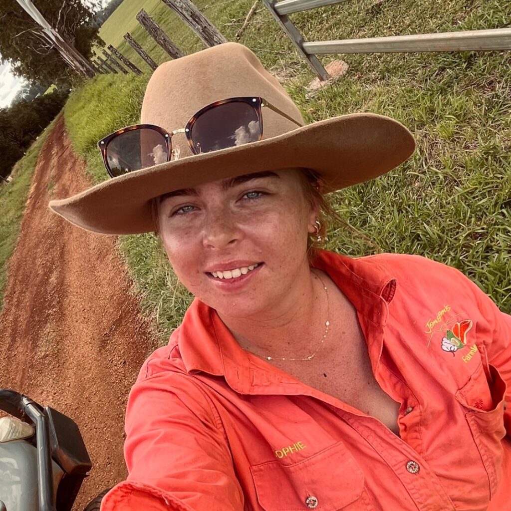 A woman wearing a wide-brimmed hat, sunglasses on top, and an orange shirt stands on a rural dirt road with a grassy fence line in the background, smiling at the camera.