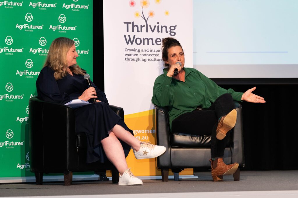 Two women sit in armchairs on a stage, speaking into microphones at the Thriving Women conference. Behind them are banners for Thriving Women and AgriFutures Australia. One woman gestures whilst talking.