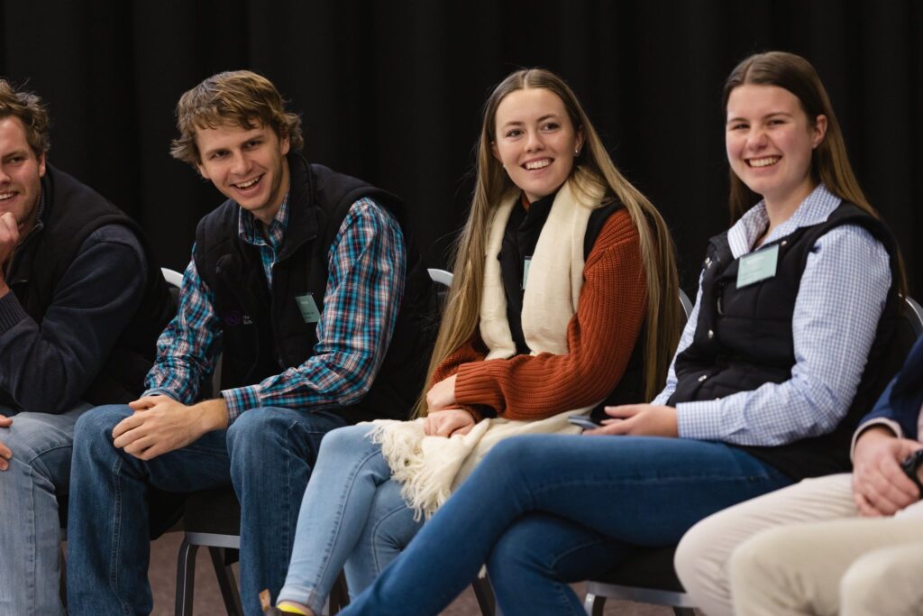 Four young adults sit in a row on chairs, smiling and engaging in conversation about the AgriFutures scholarship. They wear casual clothes, including jeans and shirts. The dark background draws focus to their animated expressions.