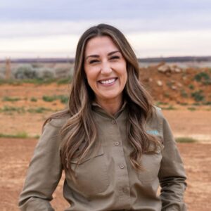 A woman with long brown hair, wearing a khaki button-up shirt, stands outdoors on a dirt landscape, smiling warmly at the camera. The background shows a cloudy sky and open land.