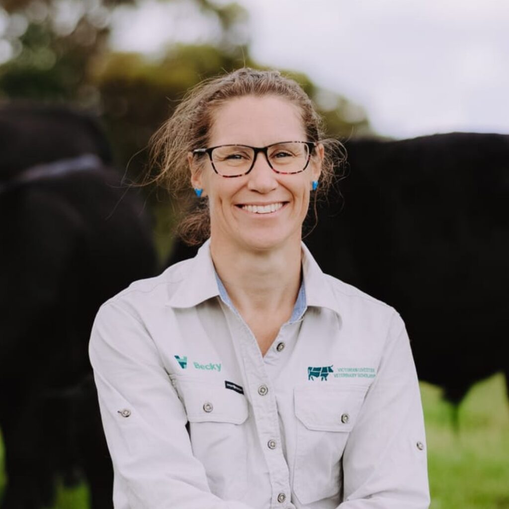 A woman wearing glasses and a light-coloured shirt with a name tag stands outside in a grassy field, smiling. Dark cows are visible in the background.