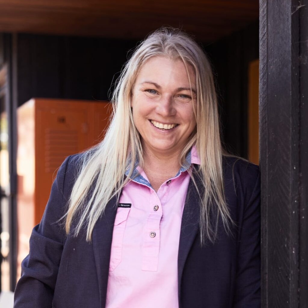 A woman with long blonde hair, wearing a light pink blouse and dark blazer, stands smiling outdoors near a building with dark walls and orange lockers in the background.