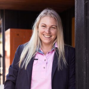 A woman with long blonde hair, wearing a light pink blouse and dark blazer, stands smiling outdoors near a building with dark walls and orange lockers in the background.