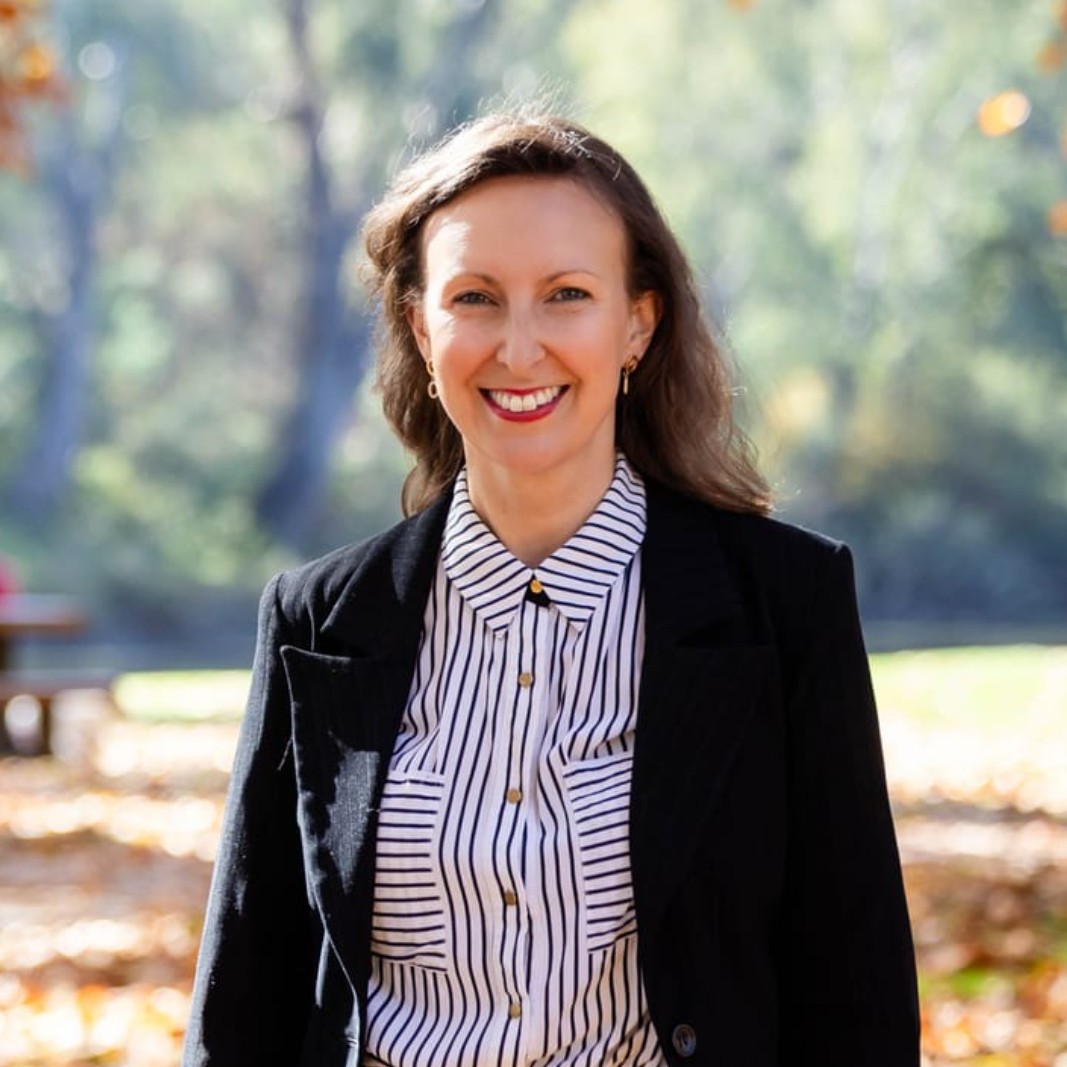 A woman with long brown hair, wearing a black blazer and a white blouse with black stripes, stands outdoors on a sunny day with autumn leaves and trees in the background, smiling at the camera.