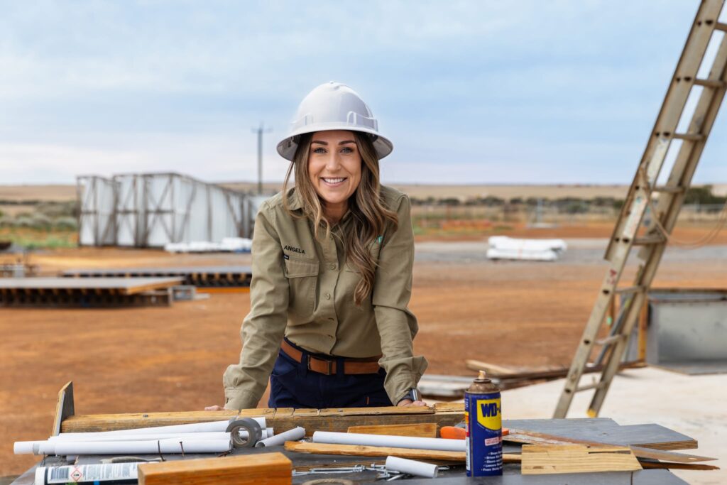 A woman in a hard hat and khaki work shirt smiles whilst leaning on a table with construction tools and plans at an outdoor worksite, embodying the spirit of regional space innovation amid brown soil and rising structures.