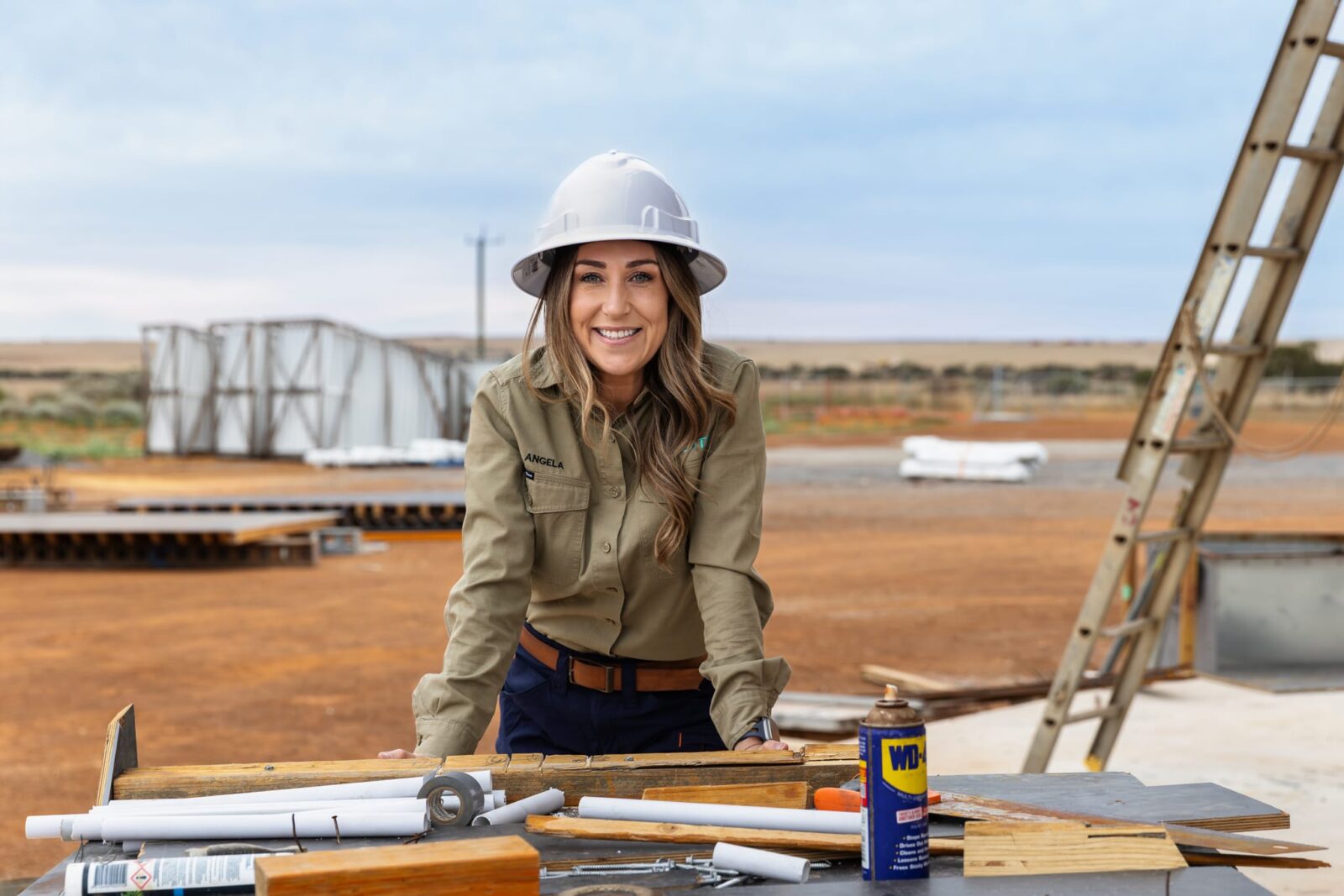 A woman in a hard hat and khaki work shirt smiles whilst leaning on a table with construction tools and plans at an outdoor worksite, embodying the spirit of regional space innovation amid brown soil and rising structures.