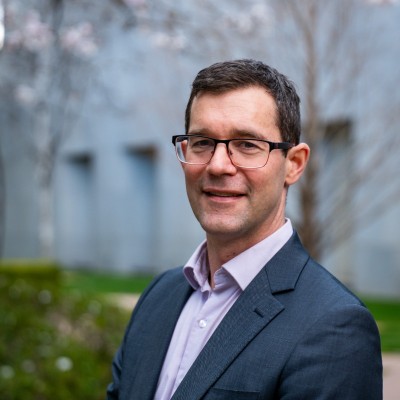 A man with short brown hair and glasses, wearing a dark suit jacket and light-coloured shirt, stands outdoors in front of a blurred background of trees and a grey building. He is smiling at the camera.