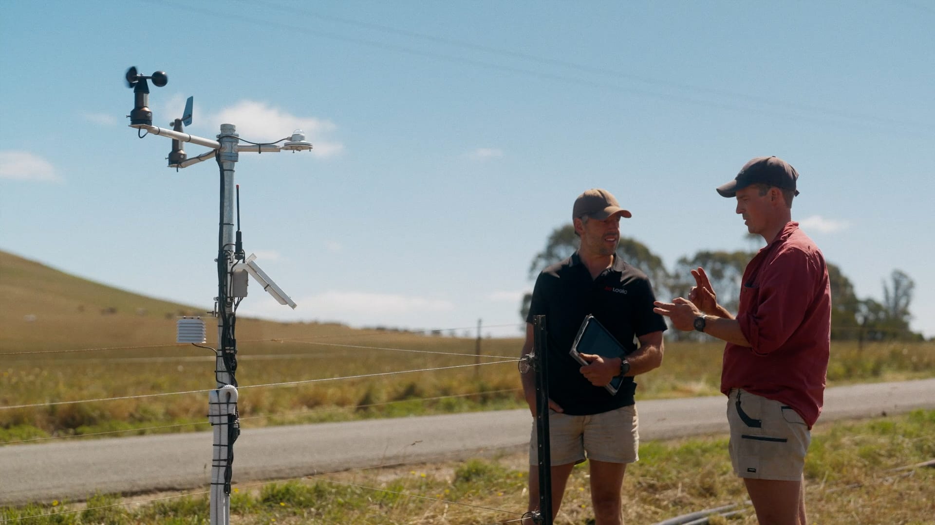Two people stand next to a weather station in a rural area, discussing data. One holds a tablet and both wear shorts, caps, and outdoor shirts. A road winds past grass, hills, and under a clear blue sky in the background.