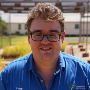A man with short, curly brown hair and glasses wears a blue shirt with CHRIS and Rice Breeding Australia embroidered on it, standing outdoors in a sunny area with buildings and plants in the background.